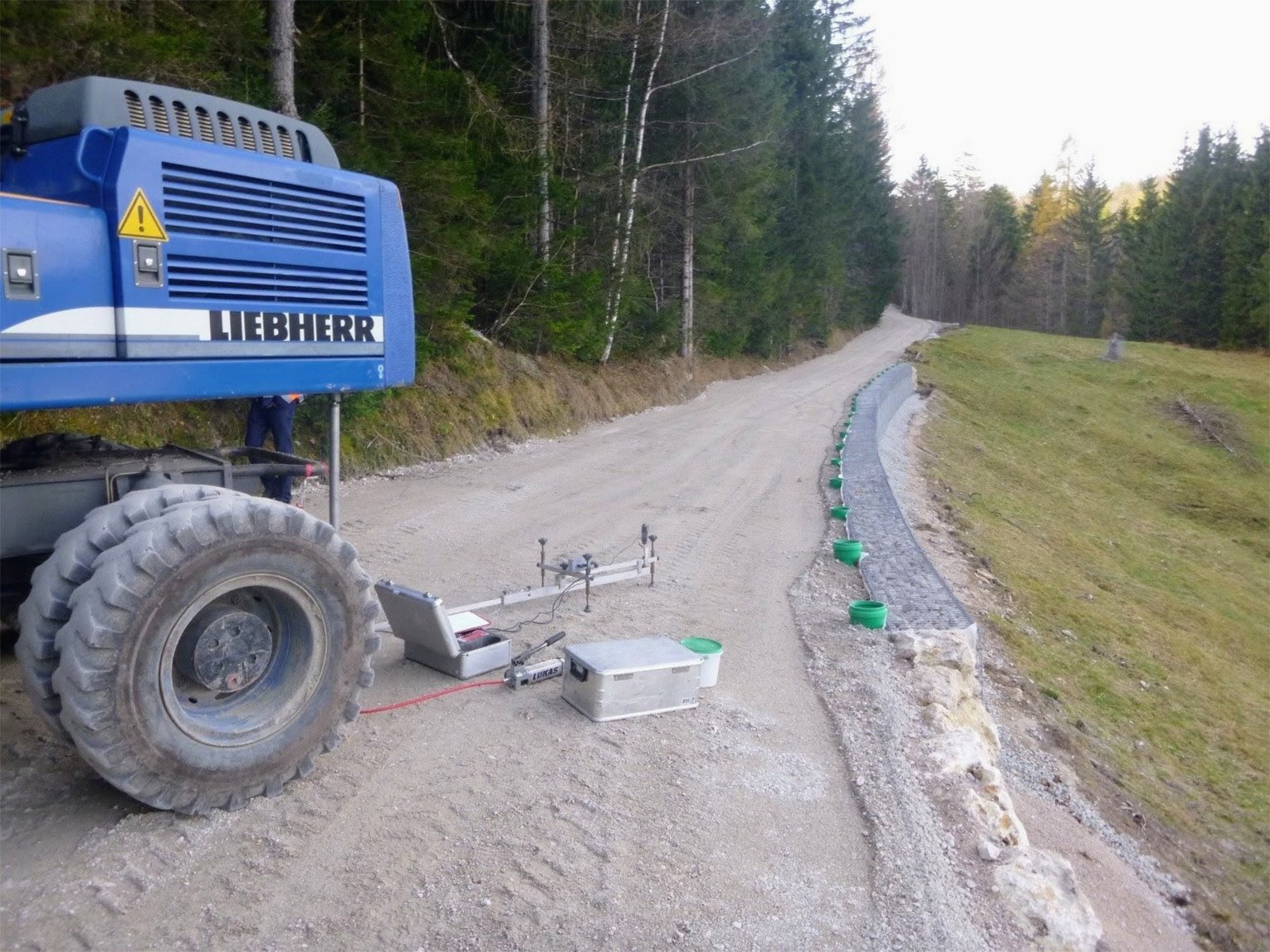 Ein blaues Bagger steht auf einer unbefestigten Straße neben einem abgesteckten Bereich, in dem GHB Consult einen statischen Plattendruckversuch durchführt.