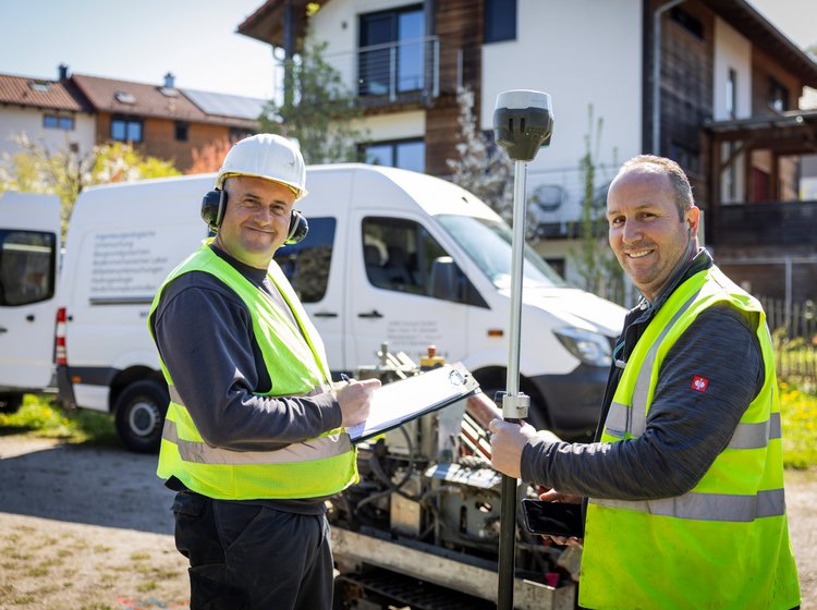 Zwei Arbeiter von GHB Consult mit Schutzweste und Helm stehen auf einer Baustelle, einer hält ein Messgerät, der andere ein Klemmbrett, im Hintergrund ein weißes Nutzfahrzeug und ein Haus.