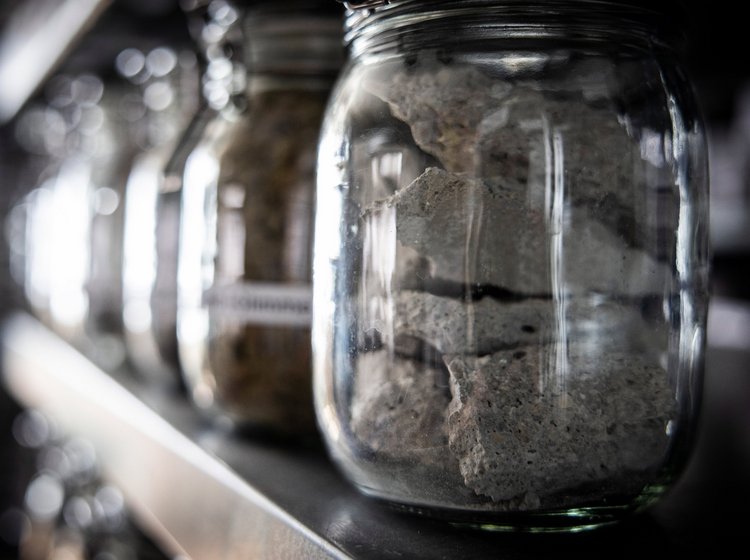 Mehrere Glasbehälter in einer Reihe, Bodenmeschanisches Labor von GHB Consult gefüllt mit verschiedenen Steinen oder Mineralien, auf einem Regal. Der Fokus liegt auf dem vorderen Glas mit hellen Gesteinsfragmenten.