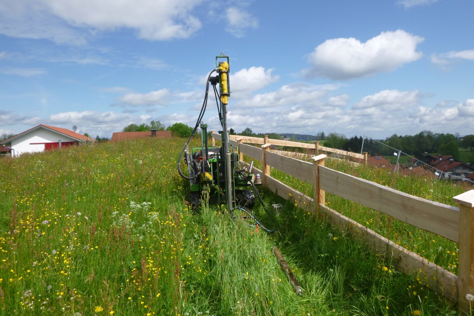 Kleinbohrung auf Wiese Bohrgerät von GHB Consult auf einer grünen, blühenden Wiese neben einem frisch errichteten Holzlattenzaun unter blauem Himmel mit vereinzelten Wolken.