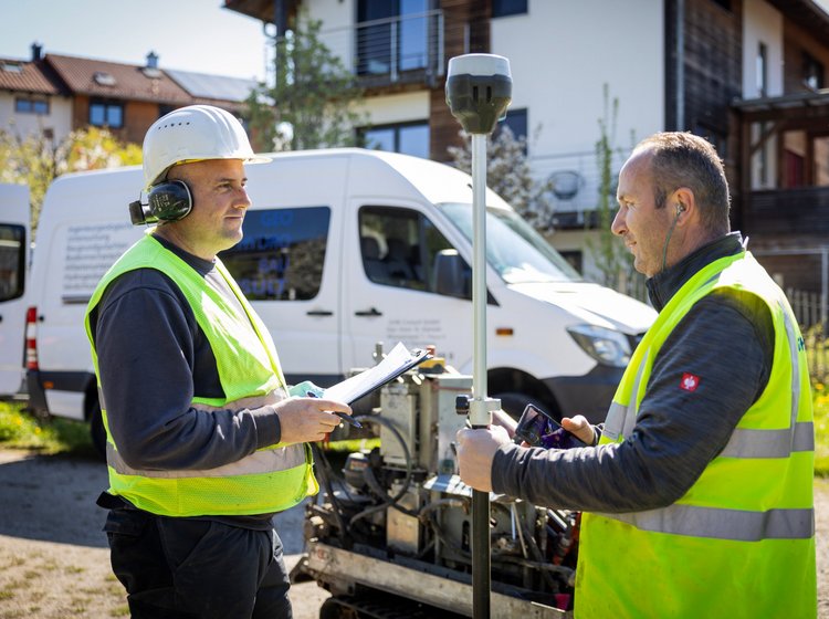 Zwei Bauarbeiter mit Schutzhelmen und Warnwesten messen im Freien mit einem Vermessungsgerät vor einem Wohnhaus.