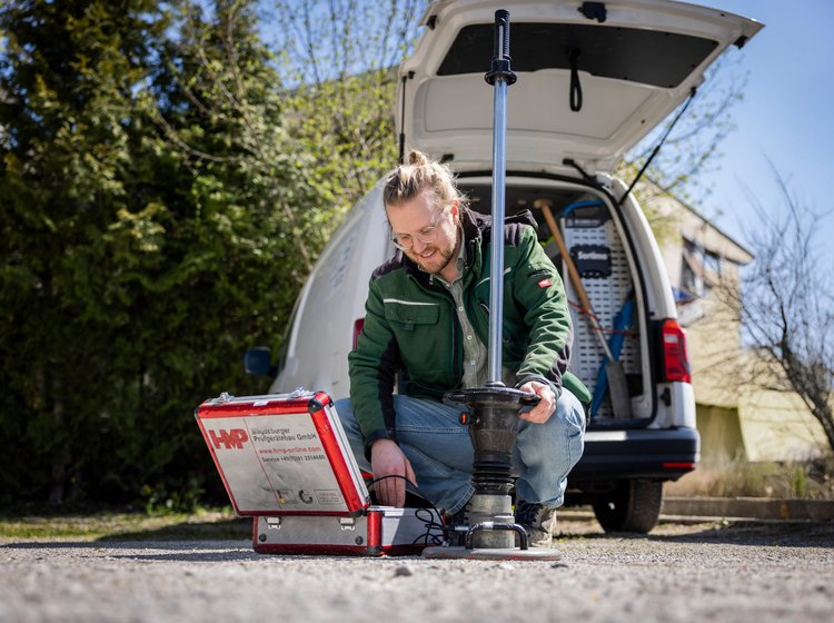 Ein Mann in grünem Jacke arbeitet im Freien mit technischem Messgerät vor einem weißen Transporter mit geöffneter Heckklappe.