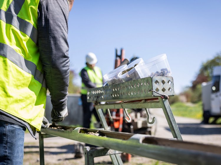 Arbeiter in Sicherheitswesten an einer Baustelle, im Vordergrund Werkzeuge und Material auf einer Arbeitsbank, im Hintergrund weitere Arbeiter und Baufahrzeuge.