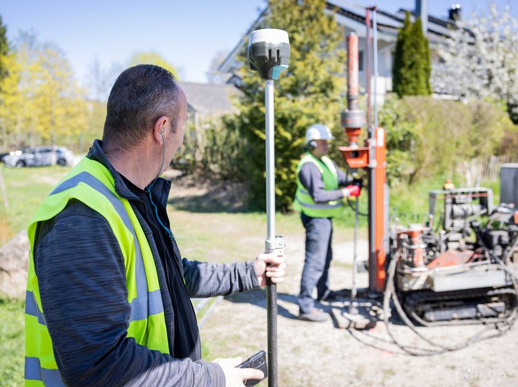 Zwei Bauarbeiter mit Sicherheitswesten arbeiten im Freien an Vermessungs- und Bohrgeräten auf einem Grundstück mit Bäumen und Gebäuden im Hintergrund.
